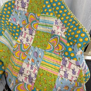 May include: A patchwork quilt featuring a variety of colourful fabric squares. Patterns include yellow circles on blue, green squares with yellow cartoon figures, striped sections in pink, yellow, and blue, and white squares with purple elephants.