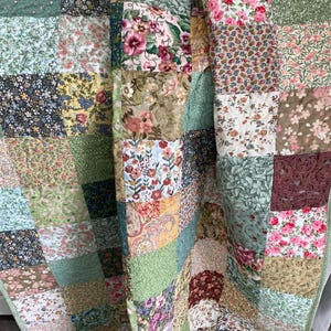 May include: A patchwork quilt featuring a variety of square fabric blocks with diverse floral, paisley, and botanical patterns in shades of green, pink, yellow, red, and blue. The quilt has visible stitching throughout and a folded edge.