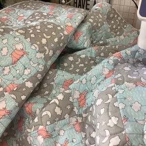 May include: A quilted fabric blanket featuring a repeating pattern of light blue and gray squares. The design includes illustrations of small sheep wearing pink pajamas, white clouds, and crescent moons, with visible machine-stitched quilting lines.
