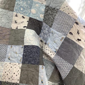 Op de afbeelding: Een patchwork quilt bestaande uit diverse blauwe, grijze en witte stoffen vierkanten. De patronen bevatten stippen, bloemmotieven en illustraties van bosdieren zoals herten, elanden en konijnen, allemaal aan elkaar genaaid met een doorgestikte textuur.