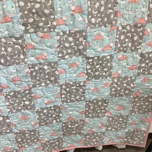 Può includere: Una trapunta patchwork con quadrati alternati di tessuto azzurro e grigio. I quadrati azzurri mostrano un motivo con agnellini rosa, mentre i quadrati grigi presentano un motivo con nuvole bianche, stelle e lune crescenti cucite insieme.