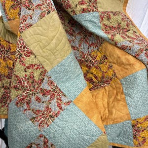 Puede incluir: Una colcha de patchwork con bloques cuadrados alternos en amarillo mostaza, verde salvia y azul claro. Los patrones de tela incluyen un estampado floral con detalles en rojo y verde, un diseño geométrico de volutas y un tejido liso texturizado.