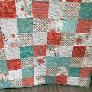 May include: A patchwork quilt featuring a repeating grid of square fabric blocks in shades of coral, teal, and white. The design includes floral patterns, geometric diamond prints, and solid-colored squares, all finished with a swirling stitch.