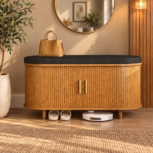 May include: A wooden storage bench with a dark gray upholstered seat and vertical slatted doors featuring two gold handles. The bench is supported by four short legs and has a tan handbag placed on top, with white slippers and a robot vacuum below.