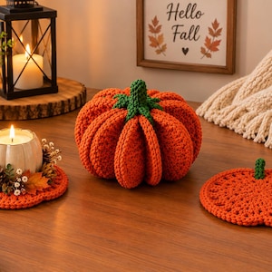 May include: A set of orange crochet items including a round pumpkin-shaped coaster, a larger three-dimensional stuffed pumpkin with a green stem, and a circular coaster base supporting a white candle surrounded by small pinecones and autumn leaves.