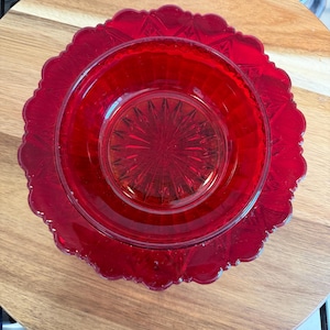 May include: A vibrant red glass bowl and matching plate set featuring a pressed glass design. The bowl has a starburst pattern at the centre with vertical ribs, while the plate has a scalloped edge and a geometric star motif around the rim.