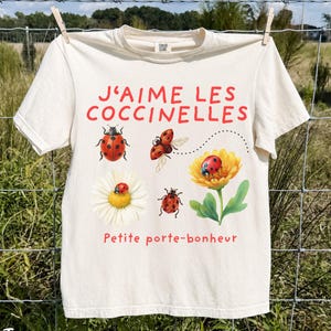 May include: An ivory short-sleeve t-shirt featuring red text that reads J'AIME LES COCCINELLES and Petite porte-bonheur. The graphic includes four ladybugs, a white daisy, and a yellow flower with green leaves, all arranged on the front.