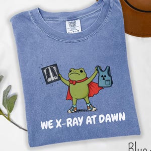 May include: A blue jean colored t-shirt featuring a graphic of a green frog wearing a red cape and sneakers. The frog holds an x-ray image and a blue vest with a radiation symbol. The text "WE X-RAY AT DAWN" is printed in white below the graphic.