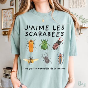 May include: A light sage green t-shirt featuring the black text J'AIME LES SCARABÉES and Une petite merveille de la nature. The design includes six detailed illustrations of various beetles in shades of orange, green, brown, and black.