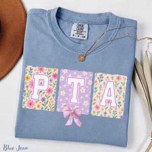 Puede incluir: Una camiseta azul de manga corta con un gráfico de las letras P, T y A en blanco con bordes rosas. Las letras están sobre fondos florales y estampados, con un lazo rosa debajo de la letra T. Un collar dorado descansa sobre el cuello de la prenda.