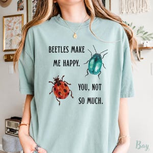 May include: A light sage green t-shirt featuring a graphic print of a red ladybird and a metallic teal beetle. The text printed in black reads, BEETLES MAKE ME HAPPY. YOU, NOT SO MUCH. The shirt has a crew neckline and short sleeves.