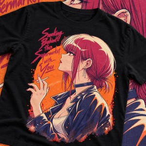 Op de afbeelding: Een zwart t-shirt met een grafische illustratie van een persoon met roodachtig roze haar in profiel, gekleed in een donker jasje en een choker. De tekst Smoking Behind the Supermarket with You staat tegen een grote oranje cirkelvormige achtergrond.
