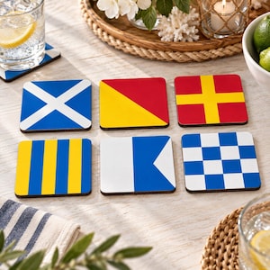 May include: A set of six square drink coasters featuring various nautical signal flags. The designs include a blue and white saltire, a yellow and red diagonal split, a red and yellow cross, yellow and blue vertical stripes, a white and blue pennant, and a blue and white checkerboard pattern.