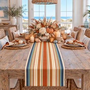May include: A long rectangular table runner featuring vertical stripes in shades of burnt orange, cream, sage green, and navy blue. The runner is centered on a rustic wooden dining table set with woven placemats, white plates, and candles.
