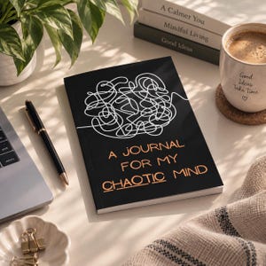May include: A black journal with a white line-art scribble design on the cover. The text "A JOURNAL FOR MY CHAOTIC MIND" is printed in a copper-colored font. The journal is rectangular with a matte finish and is positioned on a flat surface.