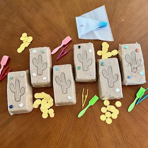 May include: Five rectangular tan blocks with embossed cactus designs, scattered with small colourful stones and gold-coloured paper circles. Includes several plastic scoops and tweezers in pink, yellow, green, and blue, plus a white piping bag.