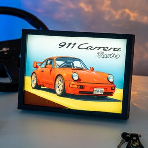 May include: A rectangular black-framed light box featuring an illustration of an orange 911 Carrera Turbo sports car on a yellow road. The text 911 Carrera turbo is printed in black above the car, which has a license plate reading BBNB-658.