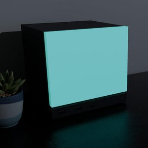 May include: A black cube-shaped electronic device featuring a large, glowing, light-blue front panel. The base of the unit includes visible ports and a circular button. The device is positioned next to a small potted succulent plant.