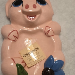以下が含まれることがあります： 光沢のある仕上げのピンク色の豚の形をしたセラミック製壁掛けプレート。青い目、笑顔の口、ペイントされた蹄が特徴です。下部には中心が暗い青い花があります。中央には「Linger Awhile」というテキストが書かれた日焼け色の紙ラベルが貼られています。