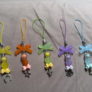 Puede incluir: Cinco colgantes para teléfono con lazos translúcidos de colores, cuentas en forma de corazón y pequeños lazos decorativos. Cada colgante cuelga de un cordón y termina con un dije de metal: una herradura, una estrella, una estrella de cuatro puntas, una flor de loto y una estrella.