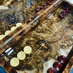Puede incluir: Un tablero de backgammon hecho de madera pulida con un patrón de vetas arremolinadas. El tablero tiene un acabado brillante e incluye un conjunto de piezas de juego circulares en colores crema y rojo oscuro dispuestas en filas sobre la superficie.