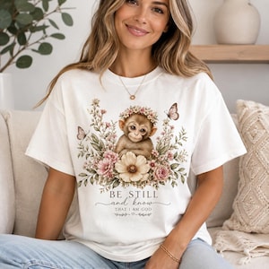 May include: A white short-sleeved t-shirt featuring a graphic of a monkey surrounded by flowers and butterflies. The text reads BE STILL and know THAT I AM GOD. The design includes soft pink, brown, and green tones on the fabric.