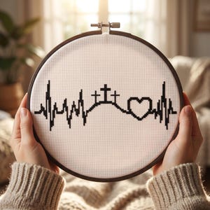 May include: A circular wooden embroidery hoop containing white fabric with a black cross-stitched design. The pattern features a continuous heartbeat line that incorporates three crosses on a hill and a heart shape in the center of the rhythm.