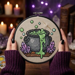 May include: A cross-stitch embroidery project in a black circular hoop featuring a black cauldron with bubbling bright green liquid spilling over the side, surrounded by purple crystals on a tan fabric background with a gold needle inserted.