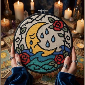 May include: A circular embroidery hoop containing a cross-stitch design of a yellow crescent moon with a sad face, blue raindrops, and red roses with green leaves. The design is set against a cream-coloured fabric background within a black frame.