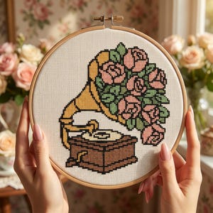 May include: A cross-stitch embroidery project in a circular wooden hoop featuring a vintage gramophone with a yellow horn. A large bouquet of pink roses with green leaves emerges from the horn. The design is stitched on white fabric.