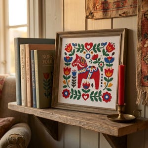 May include: A framed cross-stitch embroidery featuring a red Dala horse surrounded by a symmetrical floral pattern in red, blue, yellow, and green. The artwork is displayed on a wooden shelf next to four books and a red taper candle in a holder.