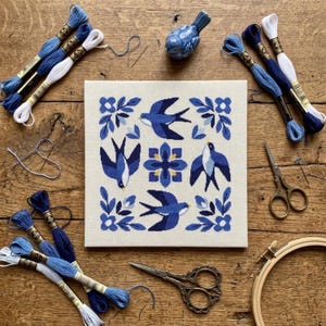 May include: A square cross-stitch embroidery piece featuring four blue birds and a central floral motif, surrounded by embroidery floss in shades of blue and white, a small ceramic bird figurine, two pairs of scissors, and a wooden hoop.