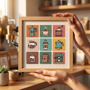 May include: A square wooden frame containing a cross-stitch sampler with nine distinct coffee-themed icons. The grid features a moka pot, a coffee cup, a grinder, a carafe, a to-go cup, a kettle, a French press, a coffee bag, and an espresso machine.