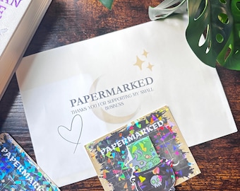 May include: A white envelope with the text PAPERMARKED and THANKS YOU FOR SUPPORTING MY SMALL BUSINESS, accompanied by two square stickers featuring a holographic cauldron graphic with a skull and the text PAPERMARKED in white lettering.