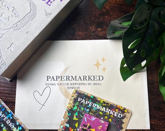 May include: A rectangular enamel pin featuring a purple background with black candle illustrations, mounted on a square card with holographic confetti accents and the text PAPERMARKED. The pin is placed near a white envelope with the text PAPERMARKED.