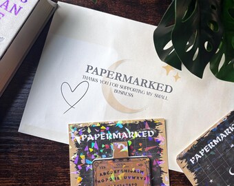May include: A rectangular pin featuring a tan ouija board design with black letters, numbers, and the words YES, NO, and GOOD BYE. The pin is attached to a square backing card with a black background, colorful holographic confetti, and the text PAPERMARKED.