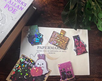 May include: A collection of five magnetic bookmarks featuring holographic designs: a cauldron with a skull, a spirit board, a haunted house, a ghost holding a pumpkin, and two candles. The bookmarks are displayed on a card with the text PAPERMARK.