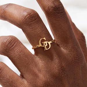 May include: A gold-toned ring featuring the letters G and D intertwined as a central focal point. The band is thin and circular, showcasing a polished metallic finish that catches the light. The design is centered on the finger, highlighting the monogram.