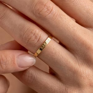 Op de afbeelding: Een dunne, gepolijste gouden ring met de naam SARAH in hoofdletters gegraveerd op de buitenkant. De ring heeft een plat, glad profiel en een metalen afwerking, wat een eenvoudig ontwerp laat zien dat duidelijk om de vinger heen zit.