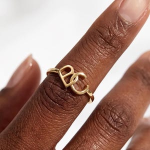 Puede incluir: Un anillo de oro que presenta dos letras mayúsculas entrelazadas, B y C, como elemento central del diseño. La banda es fina y circular, con las letras posicionadas de forma prominente en la parte superior del anillo, creando un estilo monograma.