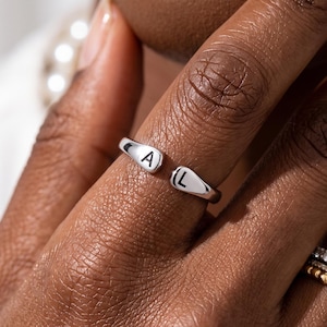 Könnte beinhalten: Ein silberfarbener offener Ring mit zwei abgerundeten Enden, die jeweils mit einem schwarzen Buchstaben graviert sind, 'A' auf der einen und 'L' auf der anderen Seite. Der Ring wird an einem Finger zusammen mit zwei weiteren Ringen getragen, einem goldfarbenen und einem silberfarbenen mit klaren Steinen.