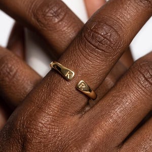 May include: A gold-toned open ring featuring two rounded ends, each engraved with a single capital letter. One end displays the letter K, while the other end displays the letter R. The band has a smooth, polished finish and a simple, minimalist design.