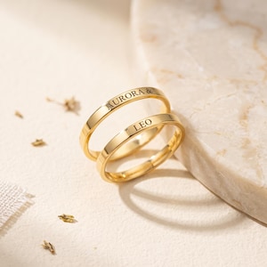Op de afbeelding: Twee gepolijste goudkleurige metalen ringen met gegraveerde tekst. De bovenste ring is gegraveerd met de naam AURORA & en de onderste ring is gegraveerd met de naam LEO. De ringen hebben een glad, afgerond bandontwerp met een reflecterende afwerking.