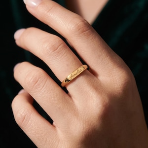 Peut inclure: Une bague dorée polie dotée d'une barre centrale plate et rectangulaire gravée du prénom Amelia dans une police cursive. L'anneau est lisse et arrondi, présentant une finition métallique qui reflète la lumière sur toute sa surface.