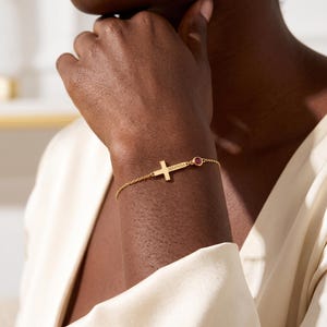 May include: A gold chain bracelet featuring a small horizontal cross charm engraved with the name Seraphina and a single round red gemstone set in a gold bezel, connected to a delicate gold link chain that wraps around the wrist.