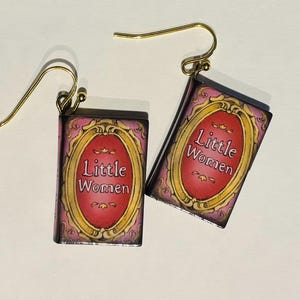 Pode incluir: Um par de brincos retangulares com design de capa de livro em miniatura. Cada brinco exibe um fundo rosa com uma moldura oval vermelha central contendo o texto Little Women em letras brancas, suspenso por ganchos de metal dourado.