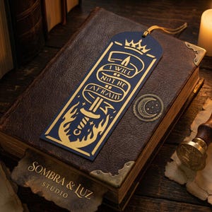 May include: A dark blue bookmark featuring a gold foil design of a sword wrapped in a ribbon with the text I WILL NOT BE AFRAID, topped with a crown and surrounded by flames, accompanied by a small circular sticker showing a crescent moon and stars.