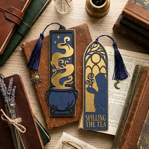 Puede incluir: Dos marcapáginas rectangulares de material azul oscuro y dorado en capas. Uno presenta un caldero con humo ascendente y un dije de sol. El otro muestra una silueta sosteniendo una taza bajo un arco con el texto "SPILLING THE TEA" y un dije de luna.