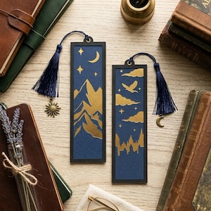 Puede incluir: Dos marcapáginas rectangulares con fondo azul oscuro y diseños en pan de oro. Uno presenta montañas, estrellas y una luna creciente con un dije de sol. El otro muestra un pájaro, nubes y la silueta de un castillo con un dije de luna. Ambos tienen borlas azul marino.