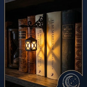 May include: A black metal bookend featuring an attached miniature lantern that emits a warm, glowing light. The lantern is suspended from a decorative scrollwork arm. The bookend is positioned between two hardcover books on a wooden shelf.